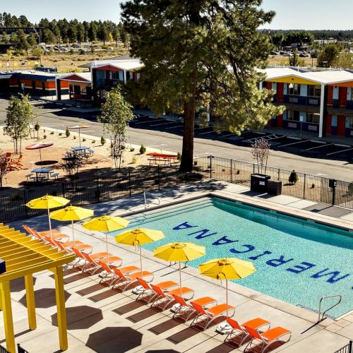 Outdoor motel courtyard with a turquoise pool, orange lounge chairs, yellow umbrellas, and apartment buildings in the background.
