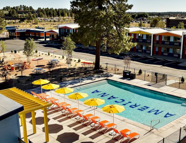 Outdoor motel courtyard with a turquoise pool, orange lounge chairs, yellow umbrellas, and apartment buildings in the background.