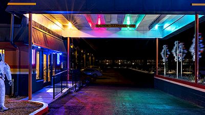 A neon-lit sci‑fi alley at night shows a person in a white spacesuit standing near a doorway under colorful lights.