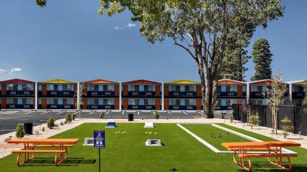 A sunny courtyard with green lawn, orange picnic benches, and a row of colorful two-story motel buildings behind a large tree.