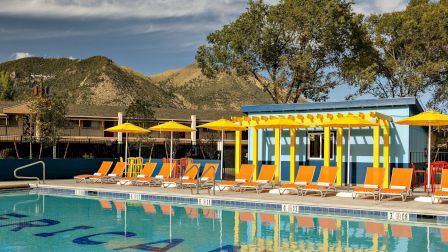 An outdoor hotel pool with orange-and-yellow loungers and umbrellas, set beside a building with mountain hills in the background.