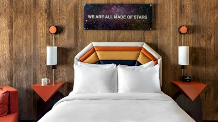 A cozy bedroom with an upholstered bed, orange bedside lamps, and a starry wall sign reading &ldquo;WE ARE ALL MADE OF STARS.&rdquo;