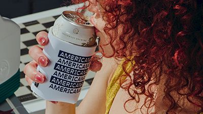 A curly red-haired woman holds a paper cup while a printer/label machine dispenses small &ldquo;AMERICAN&rdquo; cups beside her.