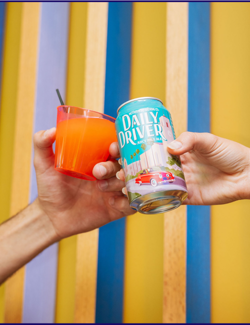 Two hands hold a Daily Driver can and orange drink in a red cup, against striped wooden boards.