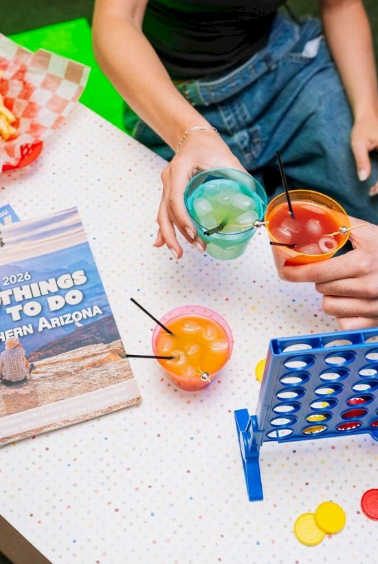 A backyard hangout with a beer-pong-style cup game, colorful balls, drinks, and a burger fries plate on a picnic blanket.