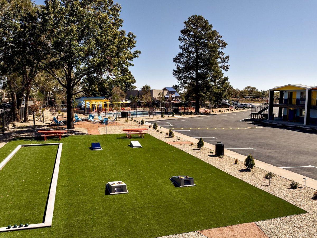 An outdoor pool complex with green lawn, benches and tables, trees, a parking lot, and colorful buildings under a clear sky.