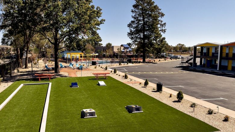 An outdoor pool complex with green lawn, benches and tables, trees, a parking lot, and colorful buildings under a clear sky.