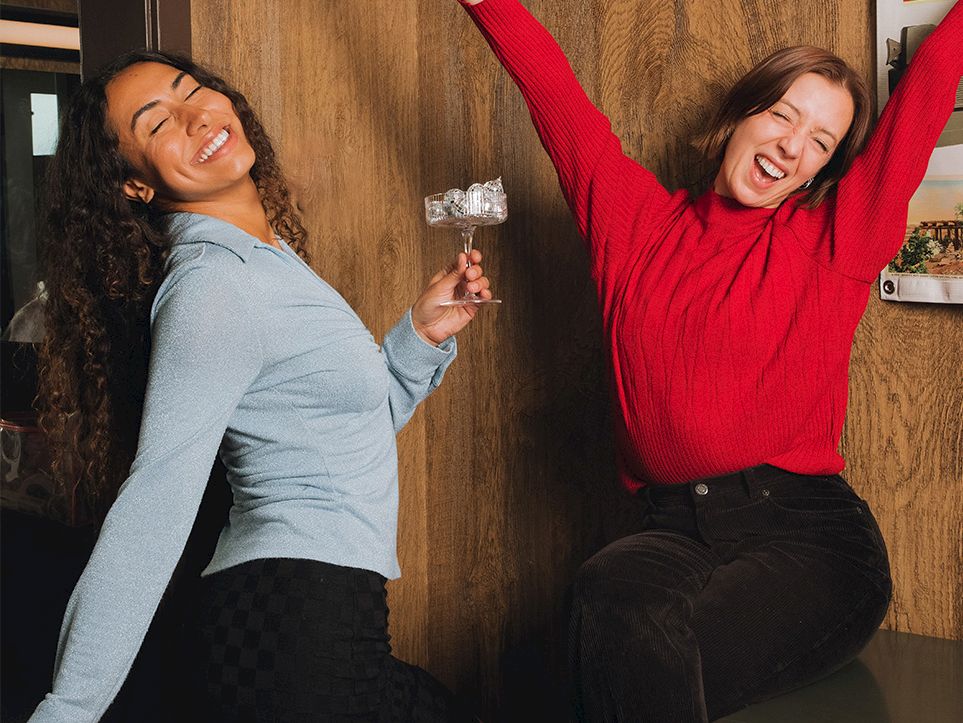 Two women sit on a couch cheering; one lifts a champagne glass while the other raises her arms, smiling.
