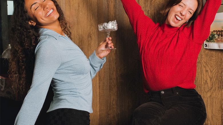 Two women sit on a couch cheering; one lifts a champagne glass while the other raises her arms, smiling.