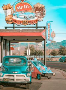 A roadside diner sign for &ldquo;Mr. D&rsquo;z&rdquo; Route 66 with burger and beer icons beside classic cars at a &ldquo;Route 66 Dinner&rdquo; stop, under a blue sky.