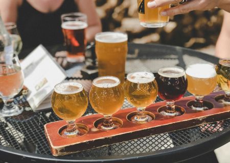 A beer tasting flight on a bar tray, with several filled glasses and a hand pouring more, at a lively brewery table.